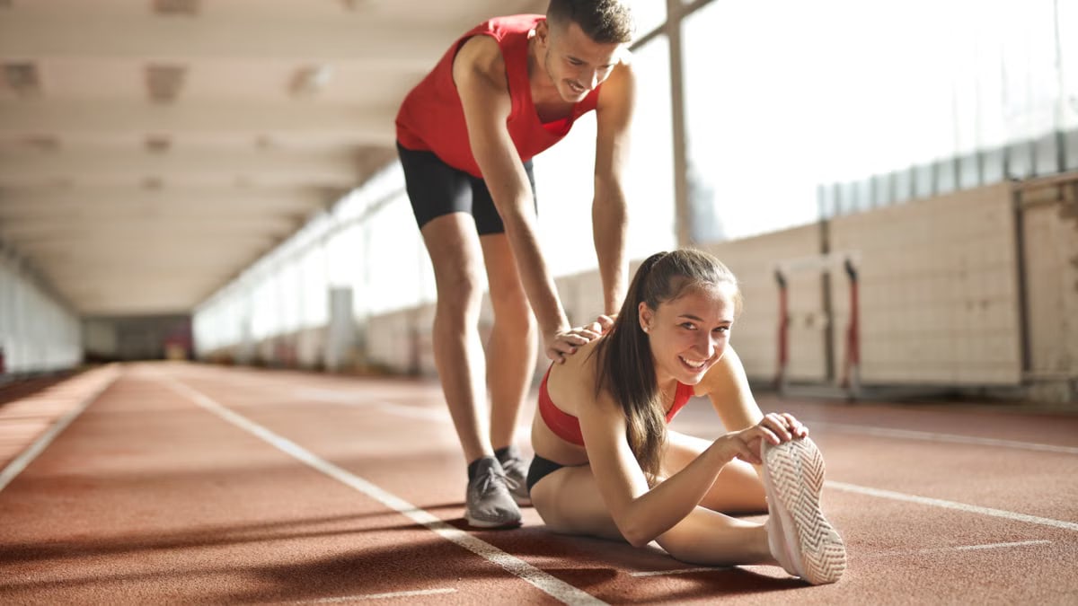 Two young athletes stretch together on an indoor track, promoting fitness and friendship.